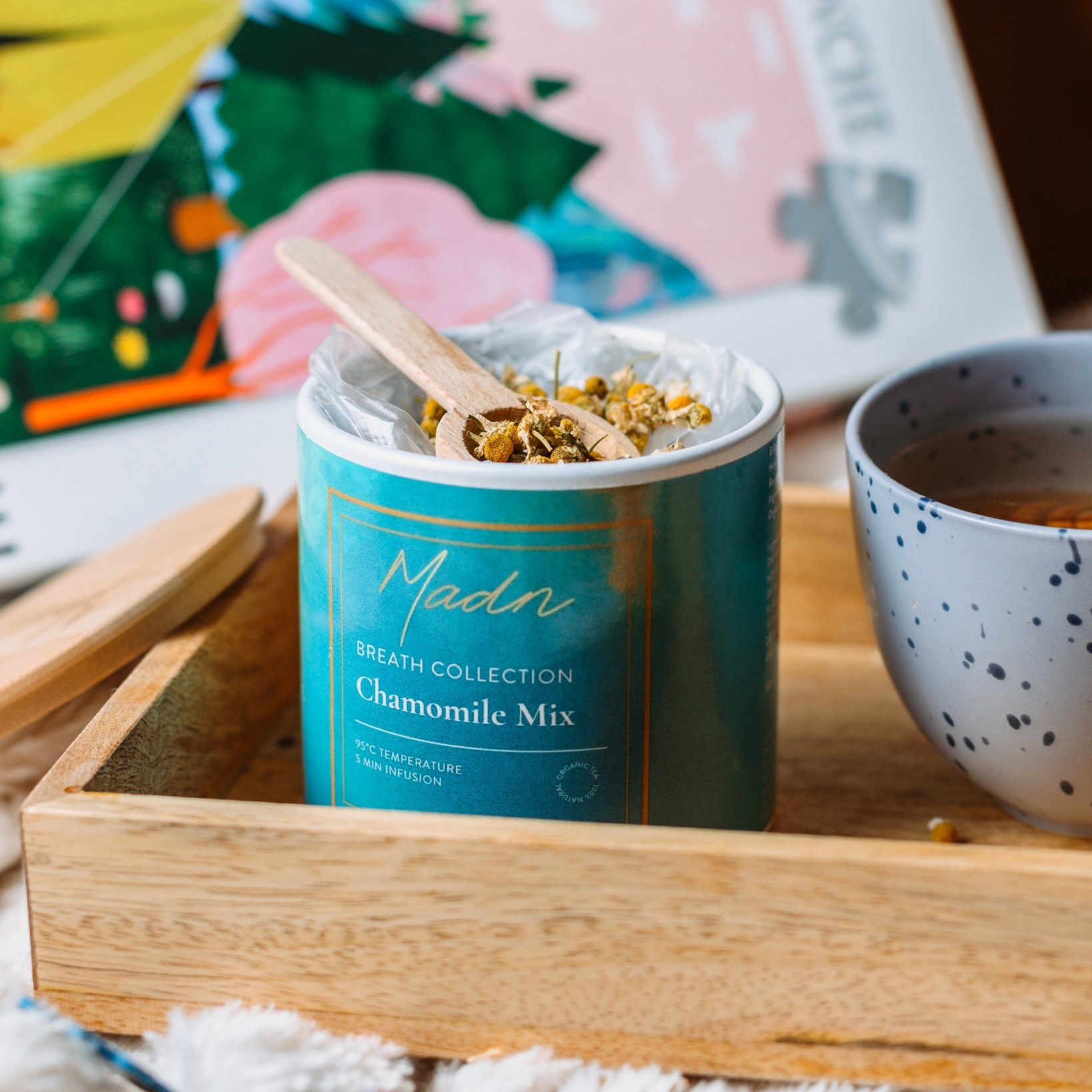 Madn “Chamomile Mix” loose-leaf tea in a teal cylinder with a wooden spoon and lid on a tray, next to a speckled cup of freshly brewed tea.
